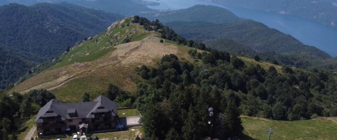 Un grande edificio borromeo si trova ai margini di una collina boscosa, affacciato su una cresta tortuosa. Sullo sfondo, un lago blu è circondato da montagne coperte di alberi, sotto un cielo limpido di kaleon.