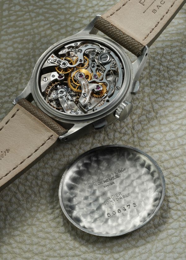 Un primo piano di un orologio da polso Patek Philippe 1518 asta aperto rivela gli intricati ingranaggi e meccanismi interni. Il fondello inciso dell'orologio, contrassegnato da testo e numeri, poggia su una superficie chiara e strutturata.
