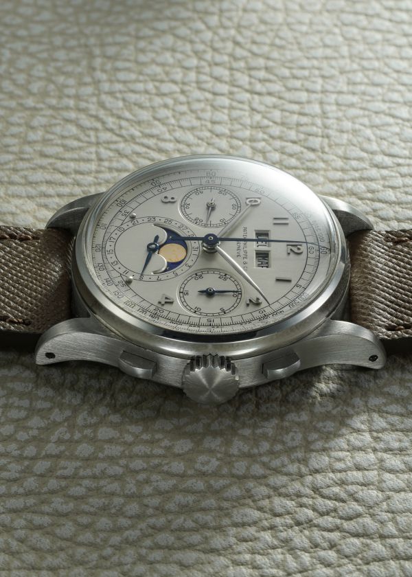 Orologio da polso d'epoca Patek Philippe 1518 asta con cinturino beige, cassa argentata e quadrante dettagliato con contatori multipli e indicazione delle fasi lunari, appoggiato su una superficie strutturata di colore chiaro.