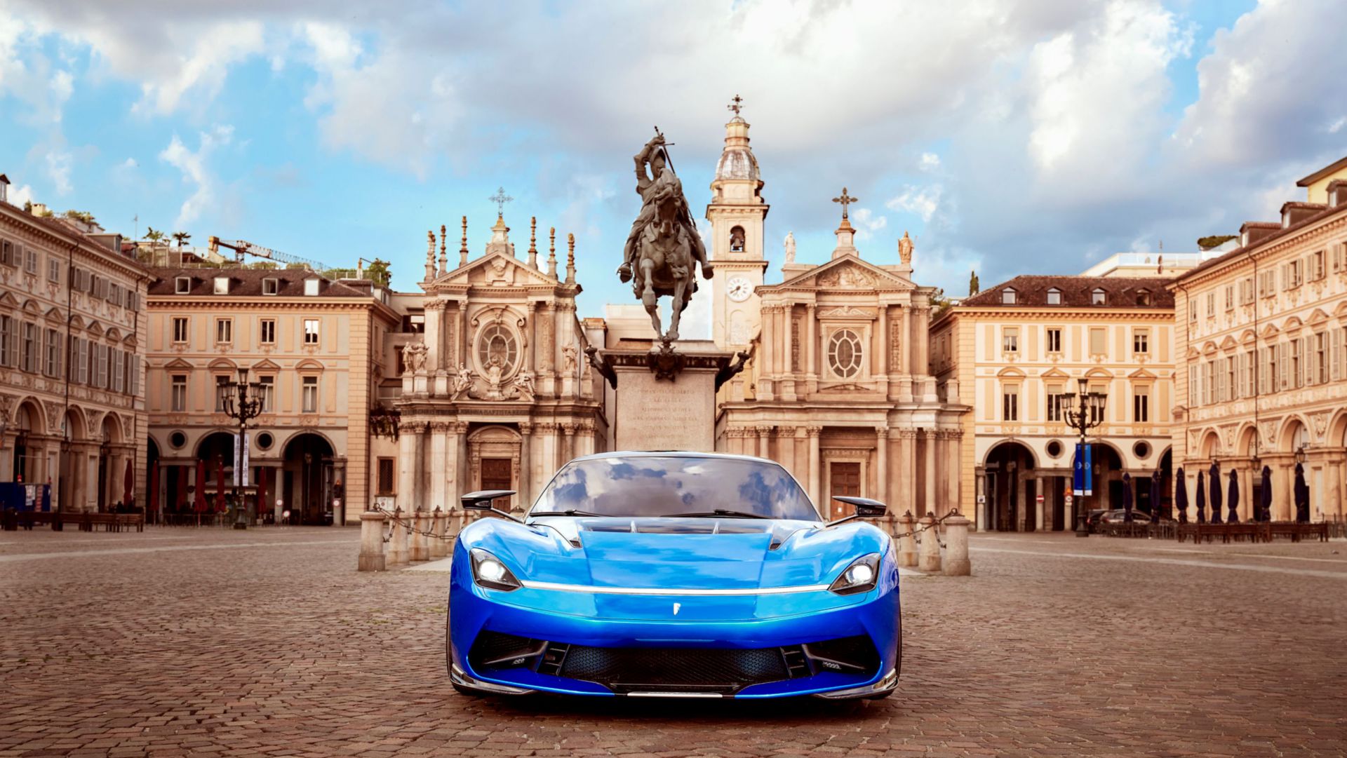 Un'auto sportiva blu è parcheggiata in una piazza acciottolata con edifici storici e una statua di cavallo e cavaliere sullo sfondo, sotto un cielo parzialmente nuvoloso.