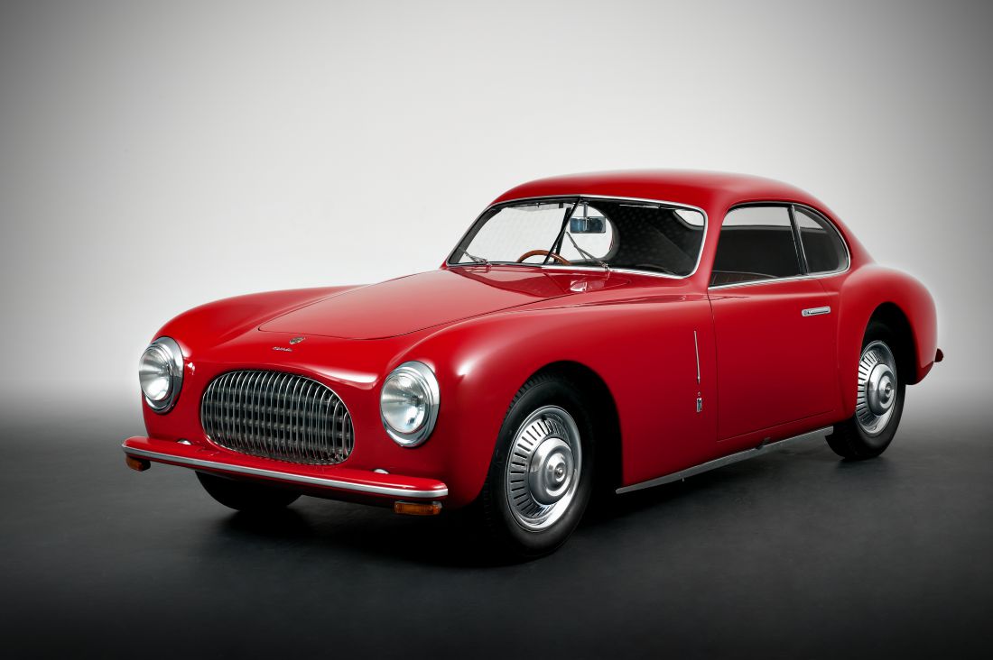Una classica auto coupé d'epoca rossa con carrozzeria arrotondata, dettagli cromati e una grande griglia anteriore - che mostra l'eleganza ispirata a Pininfarina - parcheggiata su uno sfondo grigio da studio.