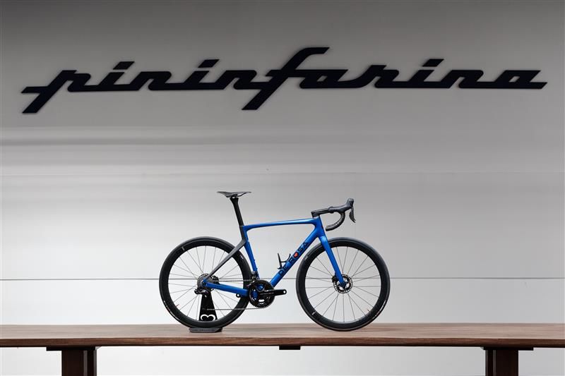 Una bicicletta da strada blu è esposta su una superficie di legno di fronte a una parete bianca, con la parola "pininfarina" scritta a grandi lettere nere in corsivo, in omaggio alla celebre casa di design italiana.
