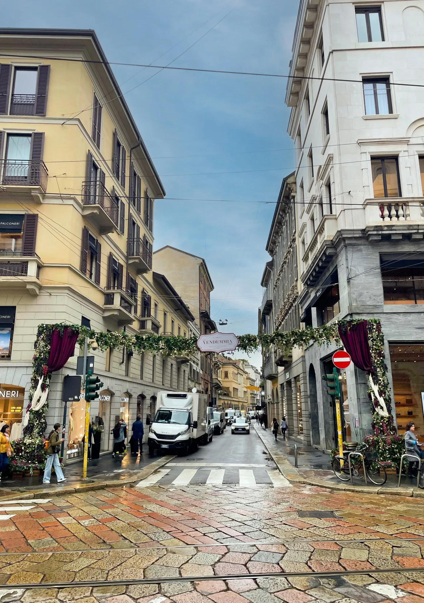 Una strada decorata in una città europea, addobbata per la Vendemmia MonteNapoleone, con festoni, alti edifici che costeggiano la strada acciottolata, pedoni che passeggiano e veicoli parcheggiati sotto un cielo nuvoloso.