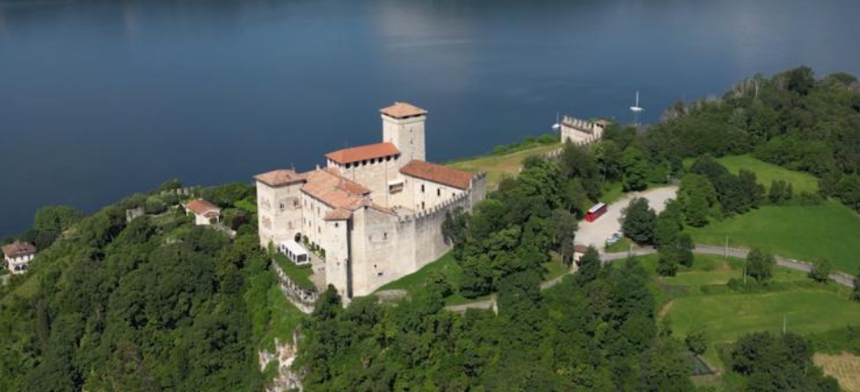 Veduta aerea del castello medievale dei Borromeo con tetti di tegole rosse, circondato da alberi verdeggianti, arroccato su una collina accanto a un grande e tranquillo lago blu.
