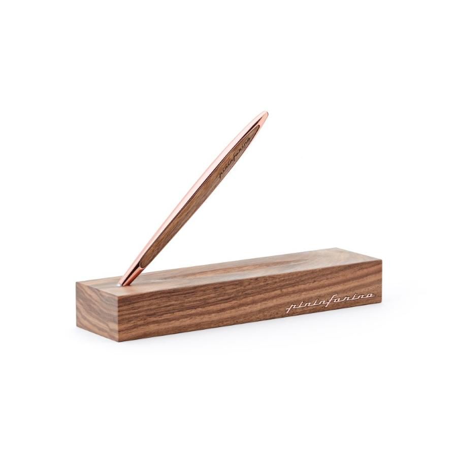 Un'elegante penna pininfarina metallizzata è bilanciata ad angolo su una base rettangolare in legno con bordi arrotondati. La base presenta un'elegante scritta bianca su un lato, creando un design minimalista e moderno.