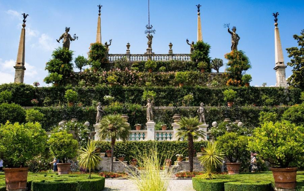 Un lussureggiante giardino italiano a terrazze ispirato all'eleganza Borromeo, con verde curato, piante in vaso e statue classiche. Colonne ornate e ringhiere decorative sovrastano il giardino in un cielo blu brillante.