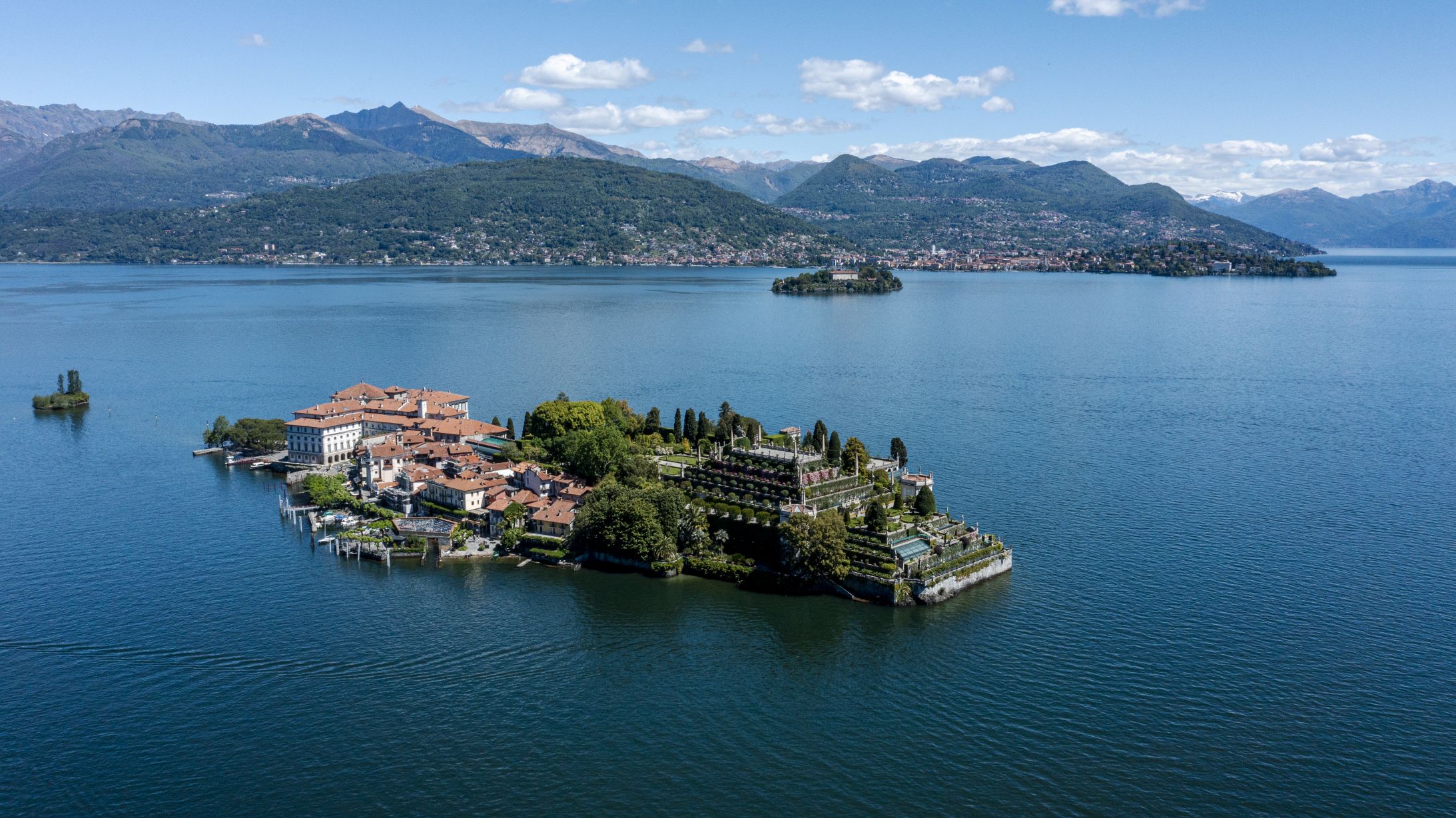Una piccola isola lussureggiante con giardini terrazzati, edifici storici Borromeo e tetti di tegole rosse si trova su un lago azzurro e tranquillo, circondato da montagne sotto un cielo parzialmente nuvoloso.