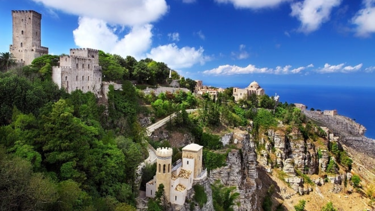 Castelli e torri medievali si trovano in cima a una collina verdeggiante a Trapani, con vista su ripide scogliere e sul mare blu sotto un cielo parzialmente nuvoloso.