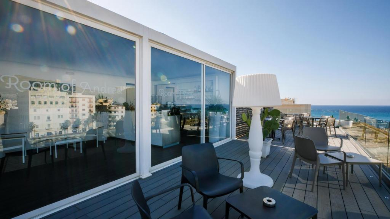 Una moderna terrazza sul tetto di Trapani con sedie nere, una lampada bianca, pareti di vetro che riflettono gli edifici della città e una vista sul mare sotto un cielo azzurro e limpido.