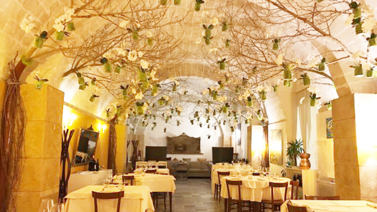 Elegante ristorante trapanese con soffitto ad arco in pietra, tovaglie bianche e rami senza foglie ornati da vasi di vetro verde e fiori bianchi sopra i tavoli da pranzo, che creano un'atmosfera calda e invitante.