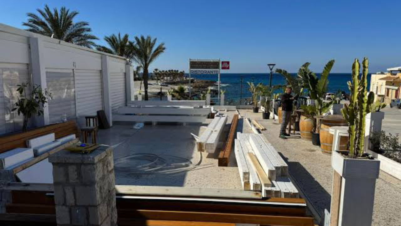 Terrazza di un ristorante sul mare a Trapani con panche e tavoli bianchi sotto un cielo azzurro e limpido. Palme, piante in vaso e due persone in piedi vicino al bordo, mentre il mare e gli edifici della città si estendono sullo sfondo.