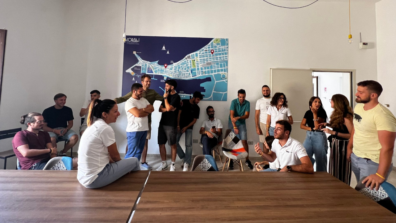 In un ufficio informale si riunisce un gruppo di persone, alcune sedute e altre in piedi, che chiacchierano e ridono insieme vicino a una grande mappa appesa alla parete che evidenzia Trapani.