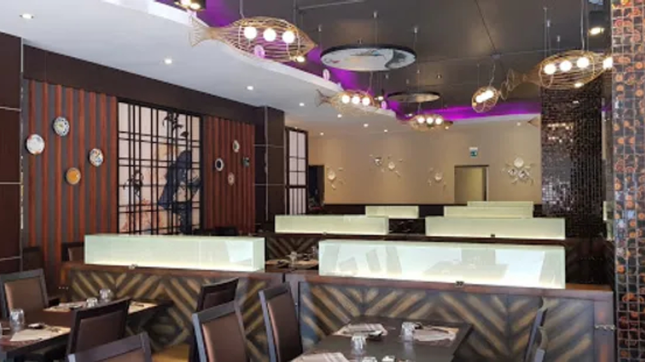 Interno di un ristorante moderno a Trapani, con cabine imbottite marroni, tavoli e sedie in legno scuro, piatti decorativi alle pareti e plafoniere a forma di pesce. L'illuminazione è calda con eleganti accenti viola sul soffitto.