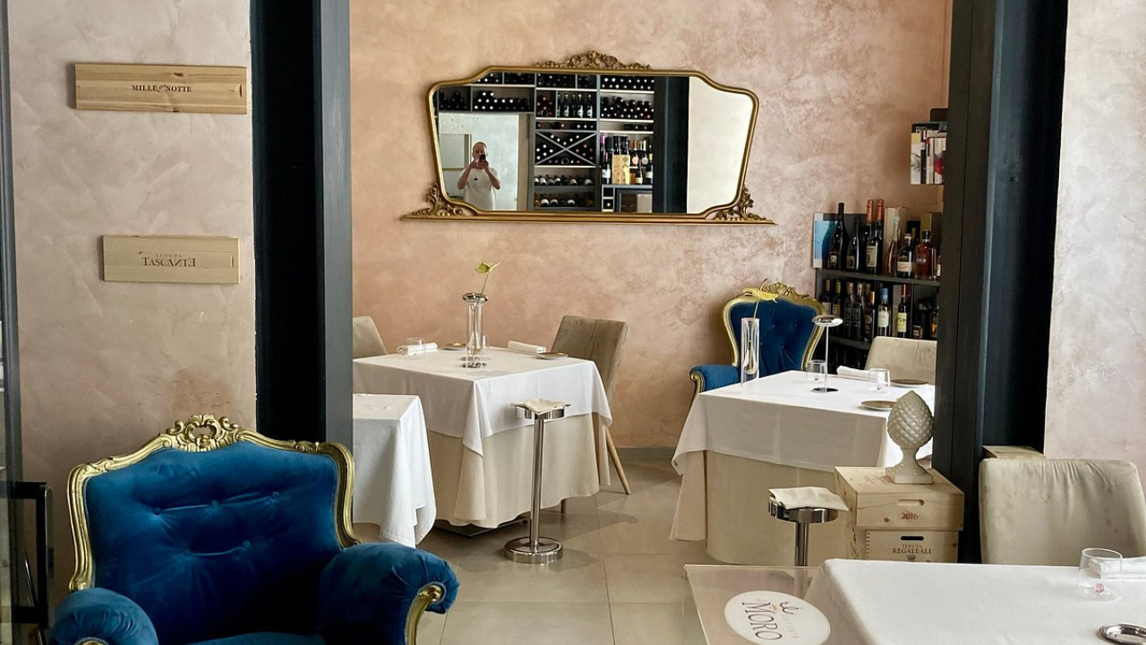 Elegante interno di un ristorante a Trapani con pareti pastello, tovaglie bianche, una poltrona di velluto blu, scaffali di bottiglie di vino e un grande specchio ornato che riflette una persona che sta scattando una foto.