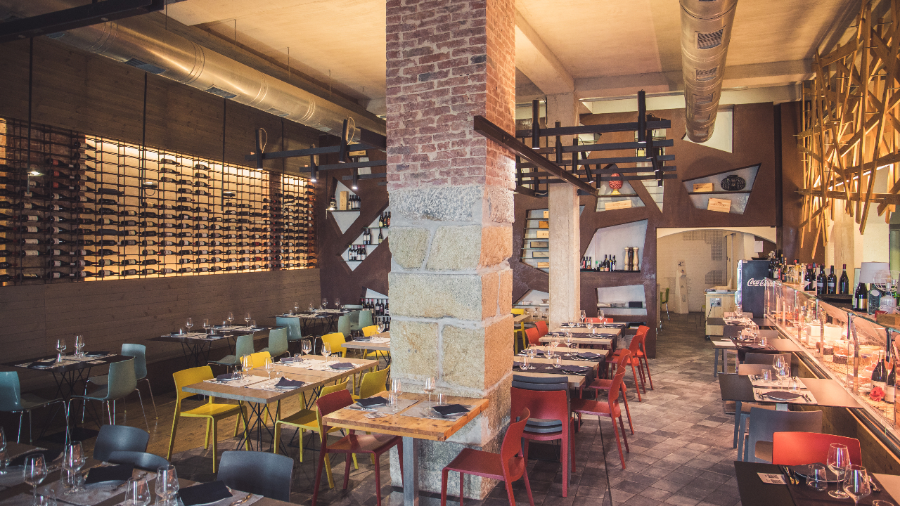 Interno moderno di un ristorante a Trapani con mattoni a vista e accenti in legno, bottiglie di vino esposte su scaffali a muro e tavoli ordinati con sedie colorate. Lo spazio è ben illuminato e invitante, pronto ad accogliere gli ospiti.