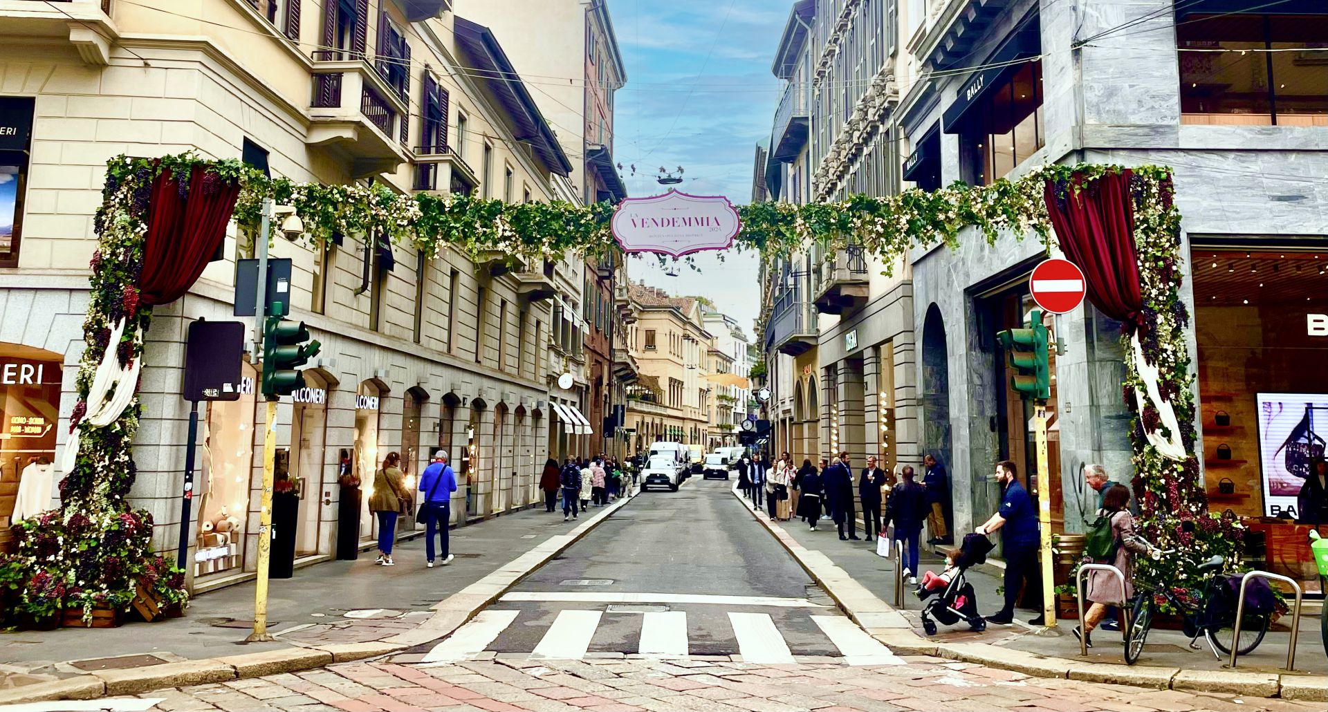 Una strada cittadina con edifici eleganti è decorata con un arco floreale e un cartello con la scritta "Vendemmia". Le persone camminano lungo i marciapiedi, alcune facendo acquisti e altre spingendo passeggini, sotto un cielo parzialmente nuvoloso.