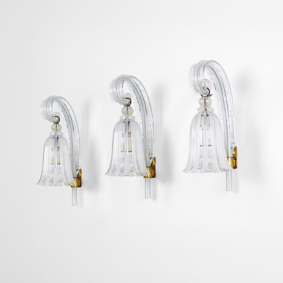 Tre eleganti appliques con bracci in vetro chiaro e curvo e paralumi in vetro a forma di campana, ciascuna montata su un supporto dorato contro una parete bianca semplice. Questo pezzo di design artistico asta settembre aggiunge un fascino delicato a qualsiasi spazio.