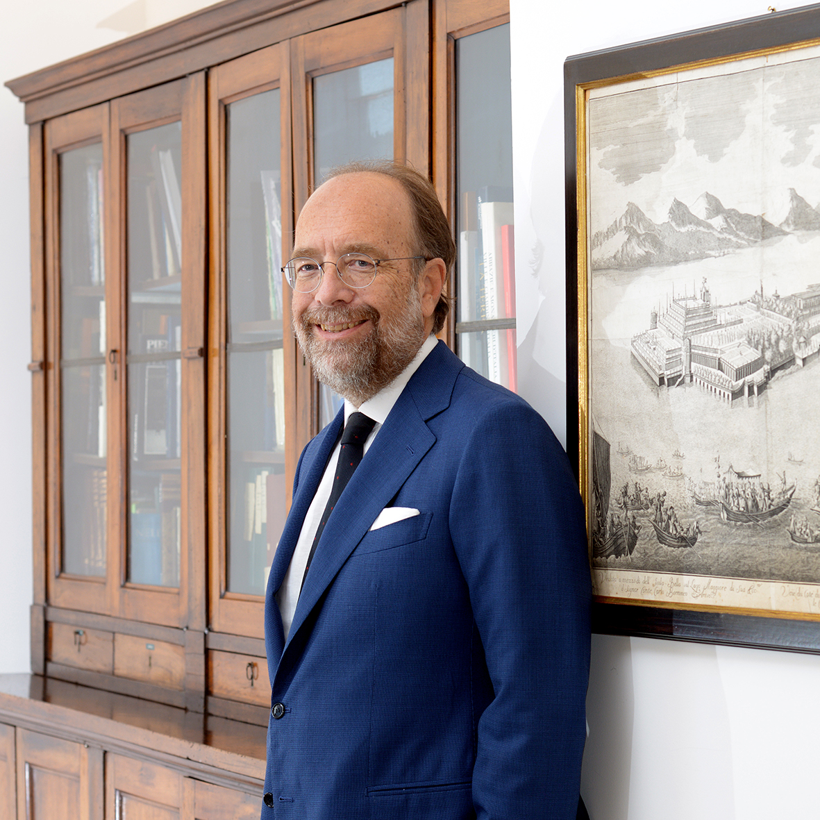 Un uomo in abito blu e occhiali sta sorridendo accanto a una libreria di legno e a un'opera d'arte in bianco e nero incorniciata che raffigura il paesaggio della città di Borromeo su una parete bianca.