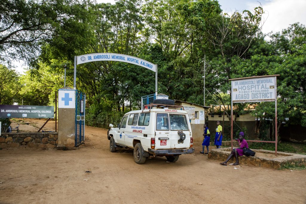 Un'ambulanza è parcheggiata all'ingresso del Dr. Ambrosoli Memorial Hospital di Kalongo, nel distretto di Agago. Sono visibili il cartello d'ingresso e il simbolo della croce, con persone e bambini nelle vicinanze e alberi sullo sfondo.