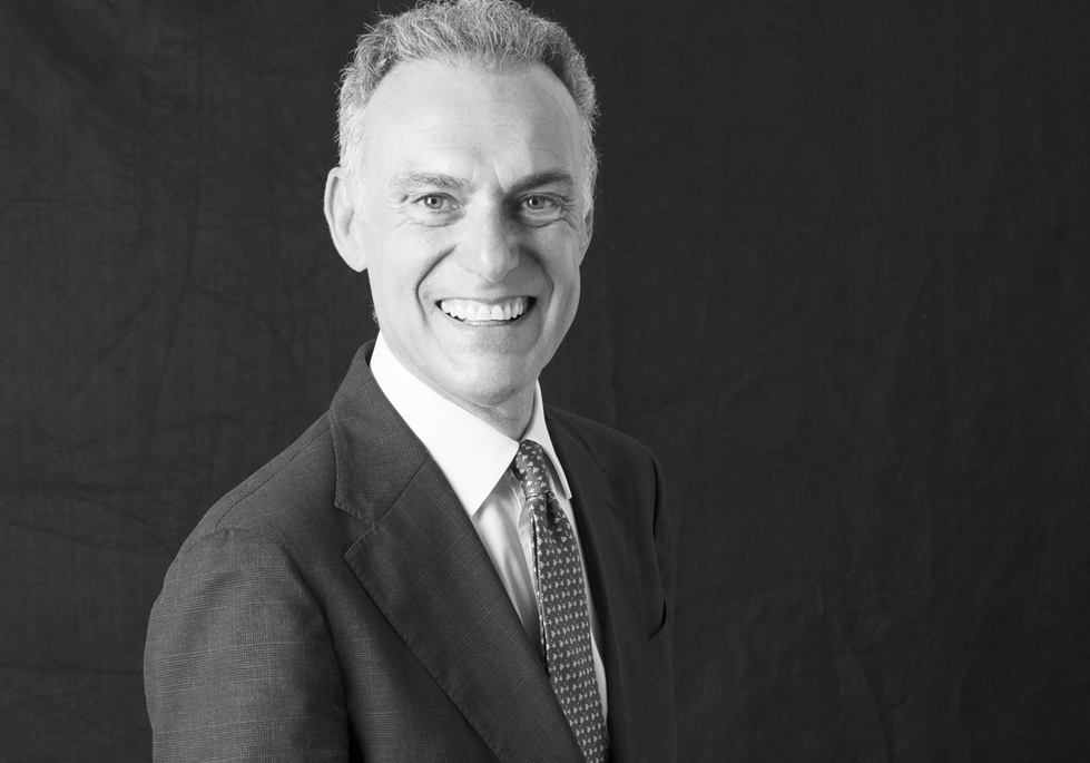 Foto in bianco e nero di un uomo sorridente in giacca e cravatta, in piedi su uno sfondo scuro e semplice. Il credito strutturato passa dagli Etf secondo Janus Henderson.