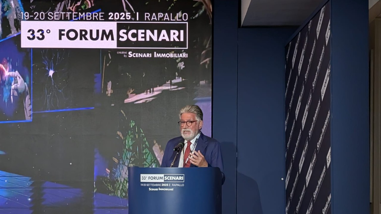 Un uomo con capelli grigi e barba parla su un podio durante il 33° Forum Scenari a Rapallo, Italia. Sullo sfondo, i dettagli dell'evento e la schermata di presentazione di un caso di studio parzialmente visibile.