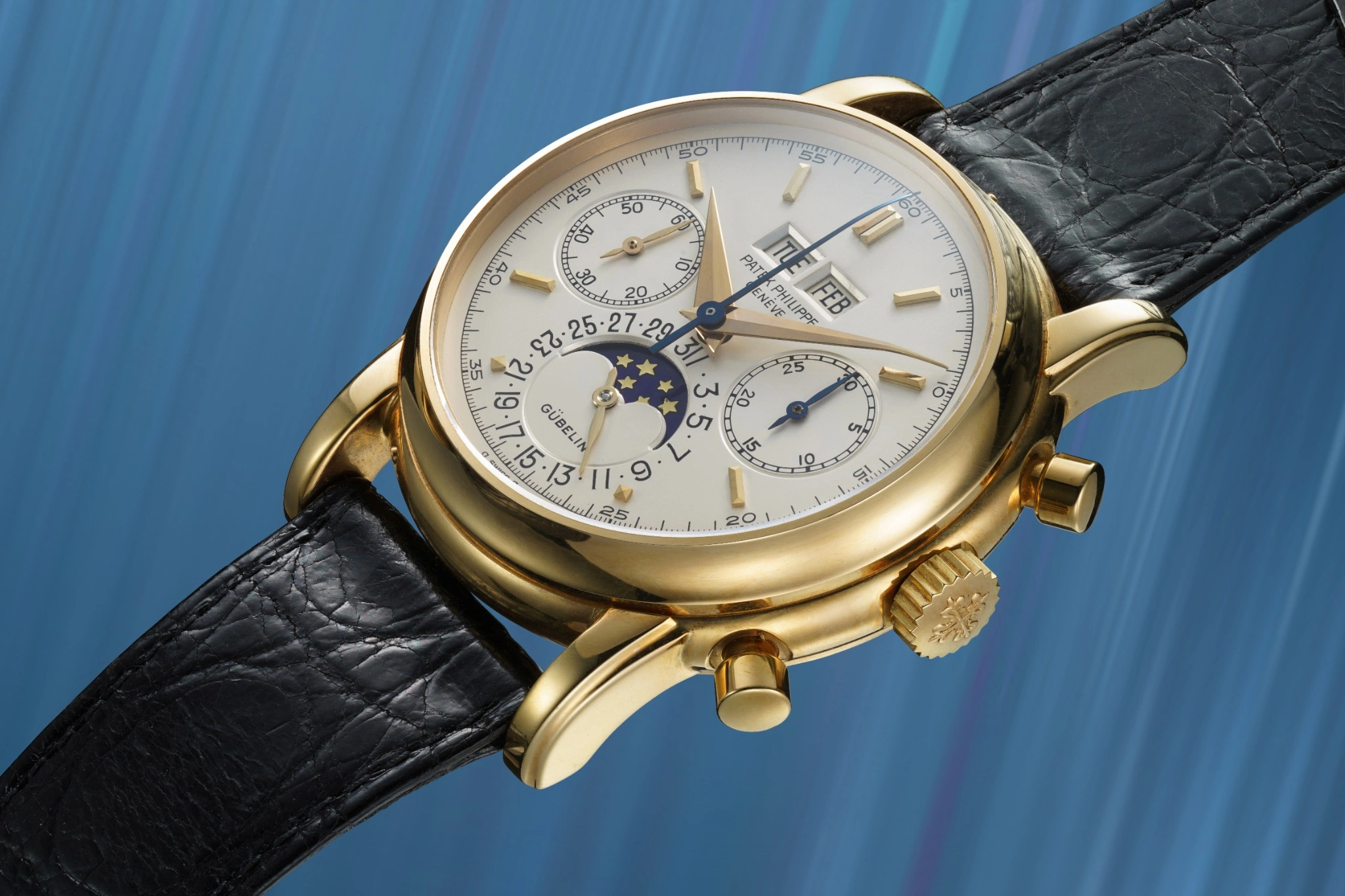Un orologio da polso in oro con cinturino in pelle nera, tre contatori, indicatore delle fasi lunari e finestrelle giorno/data su uno sfondo blu sfumato, come quelli presentati all'asta 10 anni Phillips.