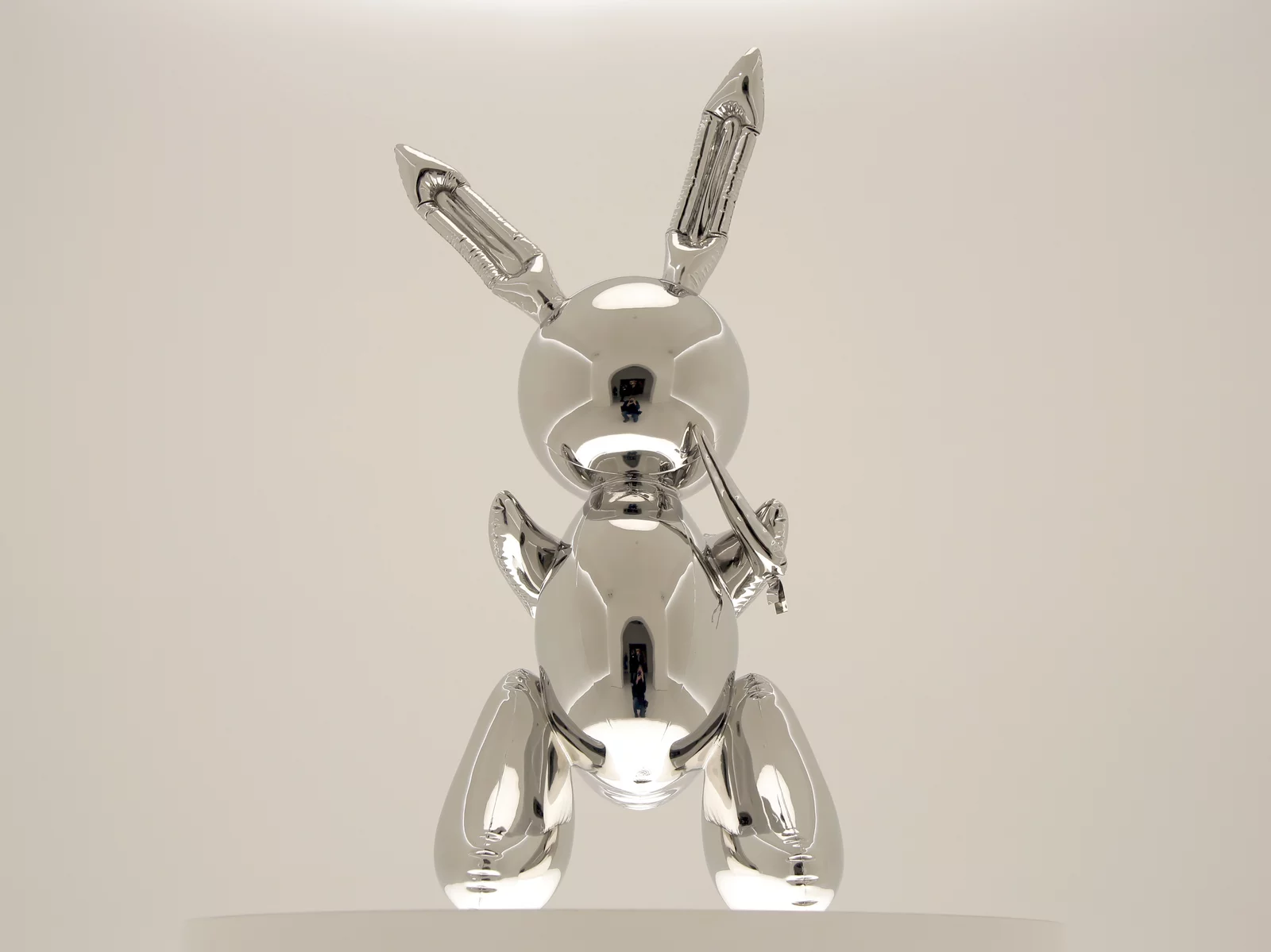 Una scultura argentata e metallica di un coniglio a palloncino che tiene in mano una carota, esposta su uno sfondo bianco su un piedistallo rotondo, richiama lo spirito giocoso delle creazioni Cattelan WC Oro.