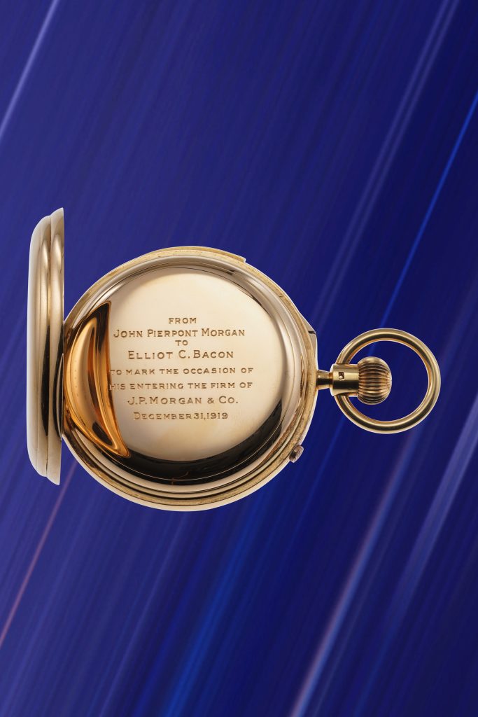 Un orologio da tasca in oro con un'iscrizione incisa è aperto su uno sfondo blu striato. Questo orologio JP Morgan commemora l'ingresso di Elliot C. Bacon nella J. P. Morgan & Co. e reca la data del 31 dicembre 1912: una rarità per qualsiasi asta.