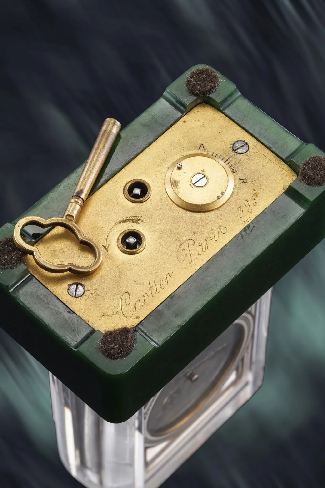 Primo piano di un meccanismo di orologio Cartier Paris d'epoca con placca in ottone e chiave di carica, incastonato in un involucro verde e trasparente. Lo sfondo, sfumato nei toni del blu scuro e del verde acqua, evoca l'eleganza degli eventi orologi JP Morgan asta.