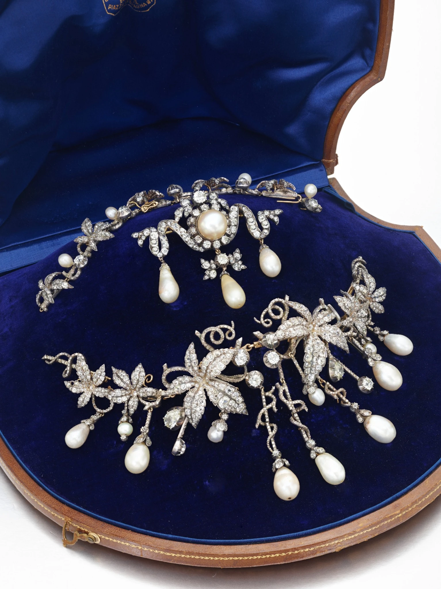 Una parure di gioielli ornati con diamanti napoleone, con una spilla e una collana di diamanti e perle in un astuccio foderato di velluto blu. Entrambi i pezzi presentano disegni floreali con perle pendenti e dettagli scintillanti.