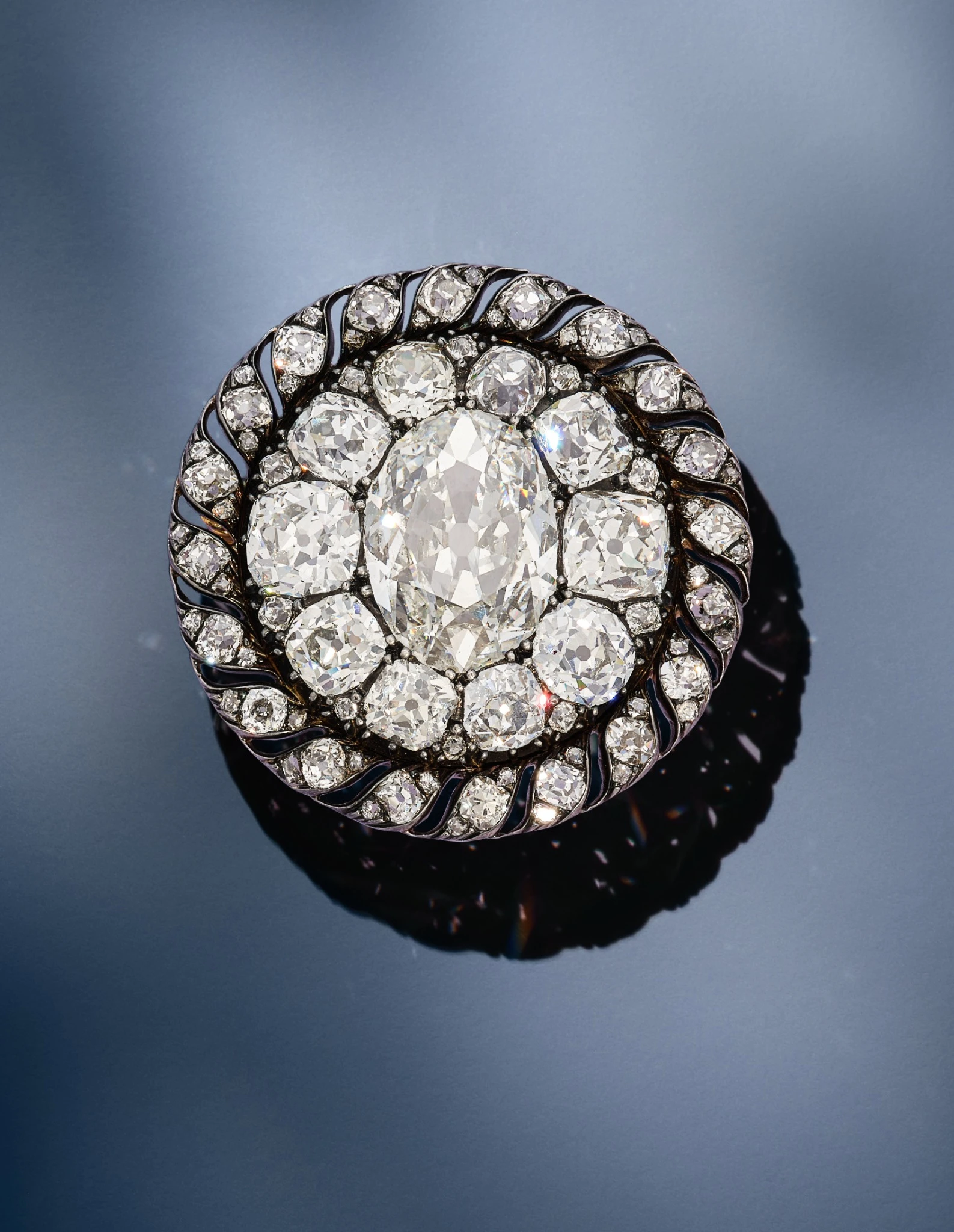 Spilla vintage rotonda con un grande diamante ovale centrale, che ricorda il famoso diamante napoleone, circondato da diamanti rotondi più piccoli in un'ornata montatura in metallo scuro, su una superficie liscia e riflettente.