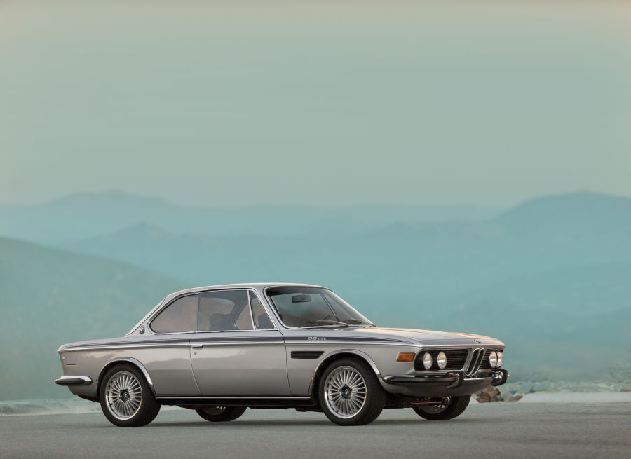 Una classica BMW 3.0 CSL coupé argentata è parcheggiata su una strada con uno sfondo di montagne azzurre e un cielo pallido.