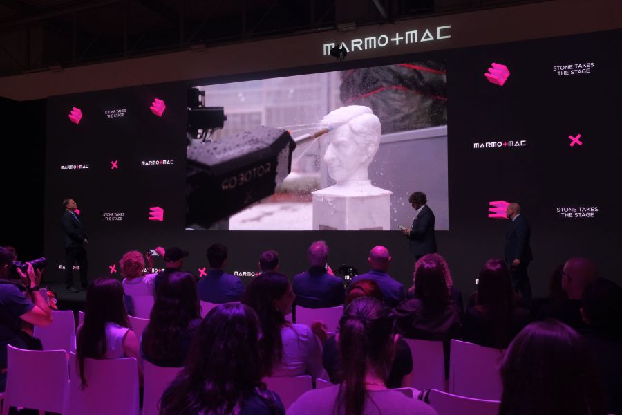 Un pubblico seduto assiste a una presentazione sul palco con tre relatori vicino a un grande schermo che mostra un braccio robotico che scolpisce un busto bianco al Marmomac. La sala, illuminata di viola e rosa, accenna all'influenza di Maurizio Cattelan sulla scena del marmo contemporaneo.