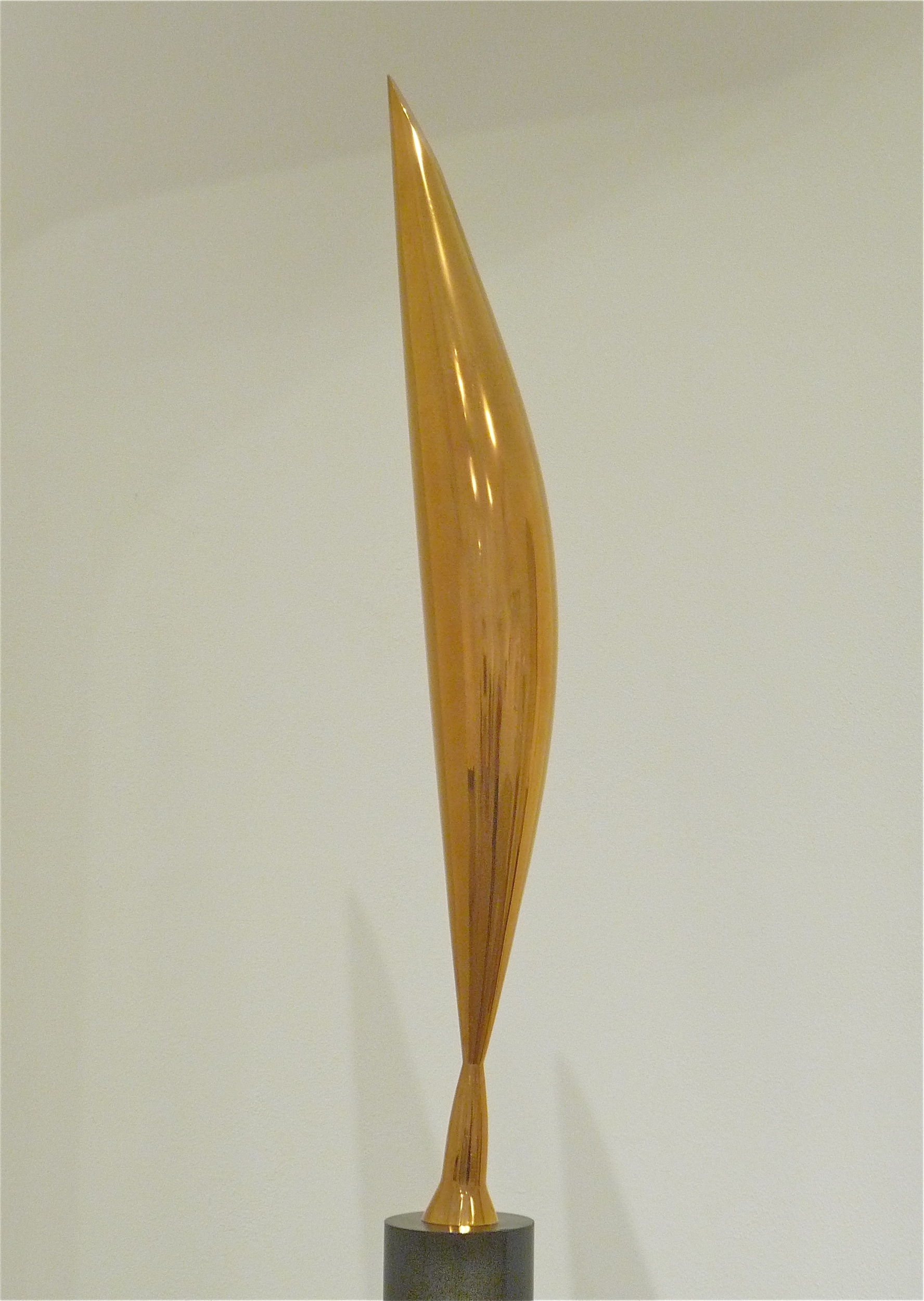 Una scultura astratta dorata, elegante e allungata, ispirata al design di Cattelan WC Oro, con una parte superiore liscia e appuntita, esposta su una base cilindrica scura su uno sfondo chiaro.