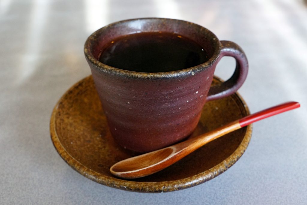 Una tazza di ceramica marrone riempita di caffè nero poggia su un piattino coordinato. Un cucchiaio di legno con manico rosso è appoggiato sul piattino accanto alla tazza. Lo sfondo è una superficie chiara leggermente sfocata.