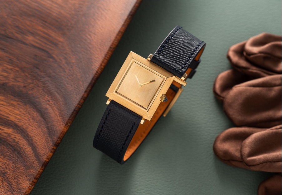 Un orologio da polso quadrato dorato con cinturino in pelle nera testurizzata, che sfoggia un raffinato design orologiero, è appoggiato su una superficie in pelle verde accanto a un oggetto in legno marrone e a un guanto marrone.