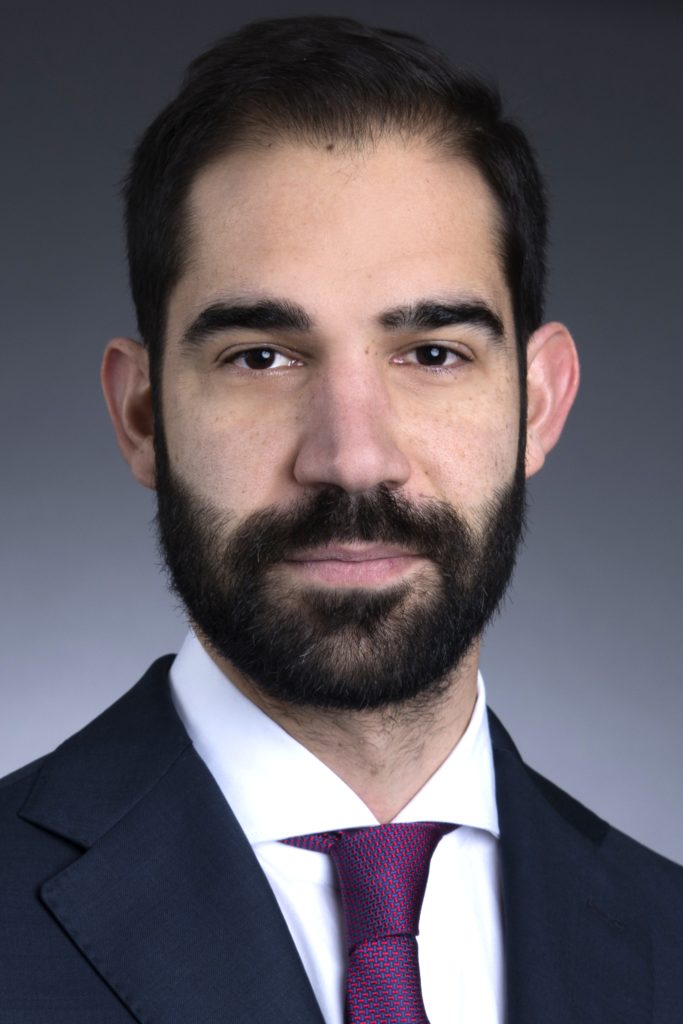 Un uomo con capelli scuri e barba folta, che indossa un abito scuro, una camicia bianca e una cravatta rossa, posa su un semplice sfondo scuro.