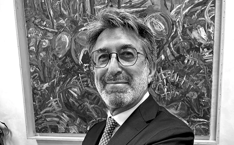 Un uomo con occhiali, barba e baffi, vestito in giacca e cravatta, sta in piedi con sicurezza davanti a un quadro astratto, un classico ritratto in bianco e nero che evoca la professionalità del mondo bancario privato.