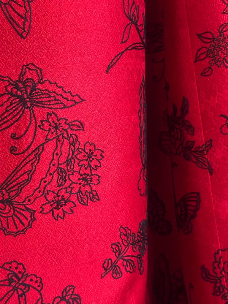 Primo piano di un tessuto rosso con un motivo strutturato, caratterizzato da disegni floreali e farfalle nere ispirati al design cinese.