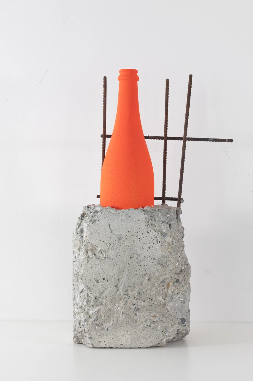 Una bottiglia di asta TOG di colore arancione brillante si trova in posizione verticale, parzialmente incastrata in un blocco di cemento grezzo con aste metalliche arrugginite che sporgono dietro di essa, il tutto su un semplice sfondo bianco.