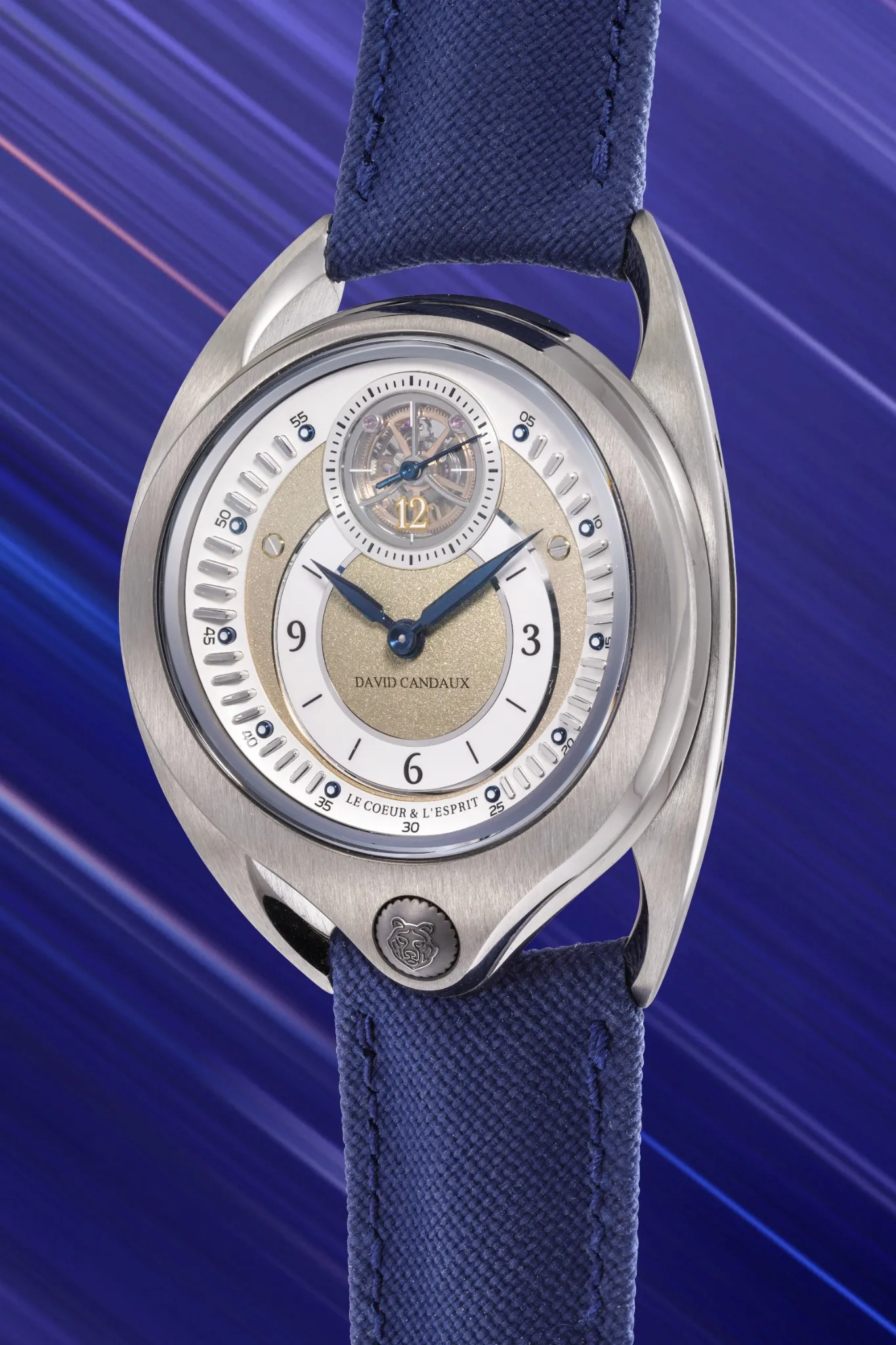 Orologio da polso di lusso della collezione prototipo con cinturino in tessuto blu, cassa argentata, quadrante dorato e argentato e tourbillon a vista a ore 12, su uno sfondo blu dinamico. Il marchio "David Candaux" è presente sul quadrante.