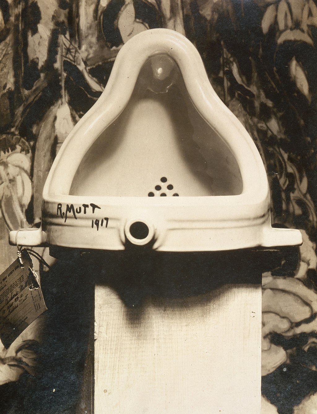 Foto in bianco e nero di un orinatoio in porcellana, che ricorda Cattelan WC Oro, posizionato a testa in giù su un piedistallo, firmato "R. Mutt 1917" sul lato con un cartellino attaccato; sullo sfondo uno sfondo a motivi astratti.