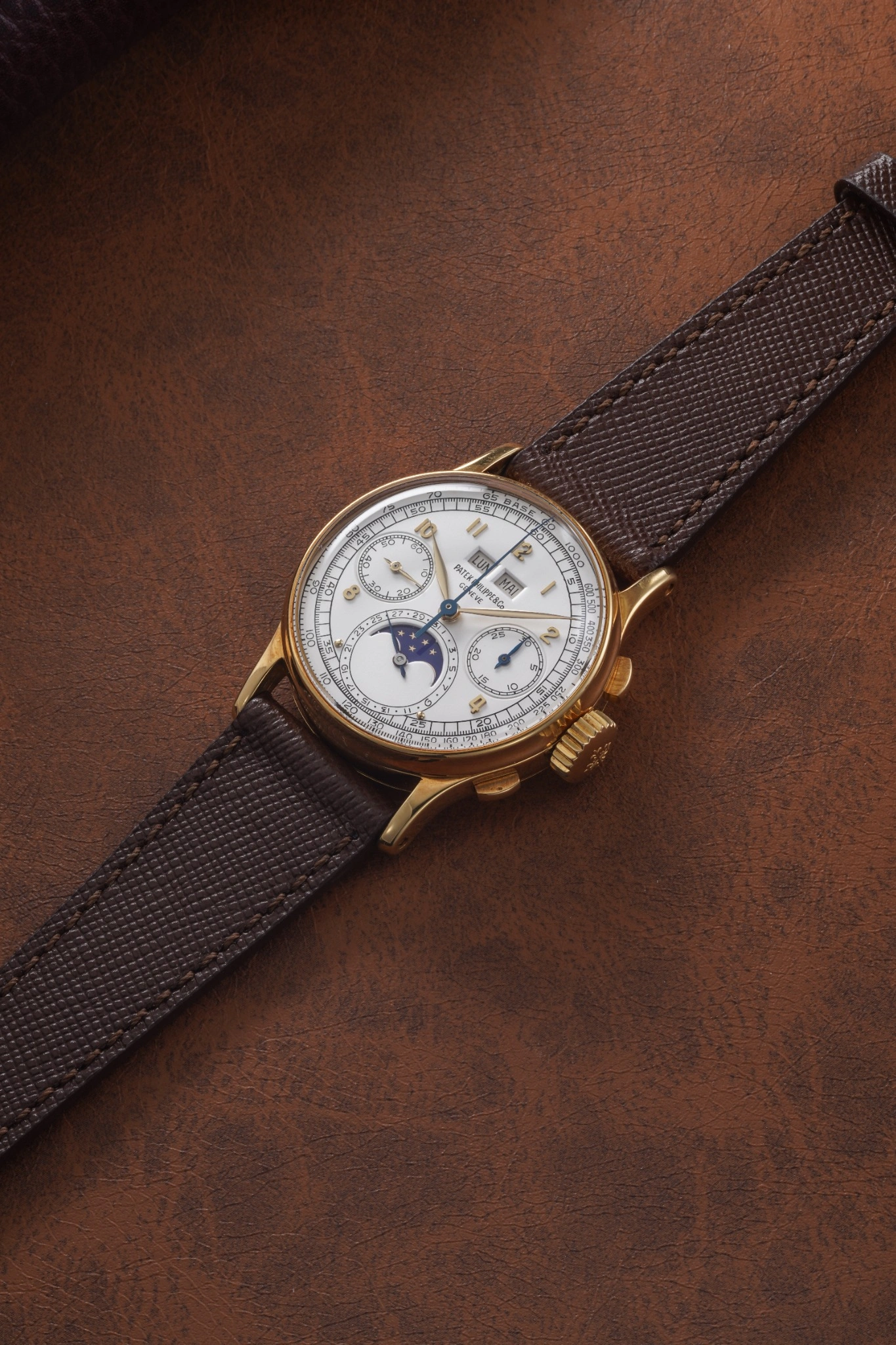 Un orologio da polso in oro con cinturino marrone poggia su una superficie in pelle marrone. Il quadrante dell'orologio presenta più quadranti - fasi lunari, giorno, data e indicatori del mese - un pezzo senza tempo che ricorda i pezzi forti delle aste 10 anni Phillips.