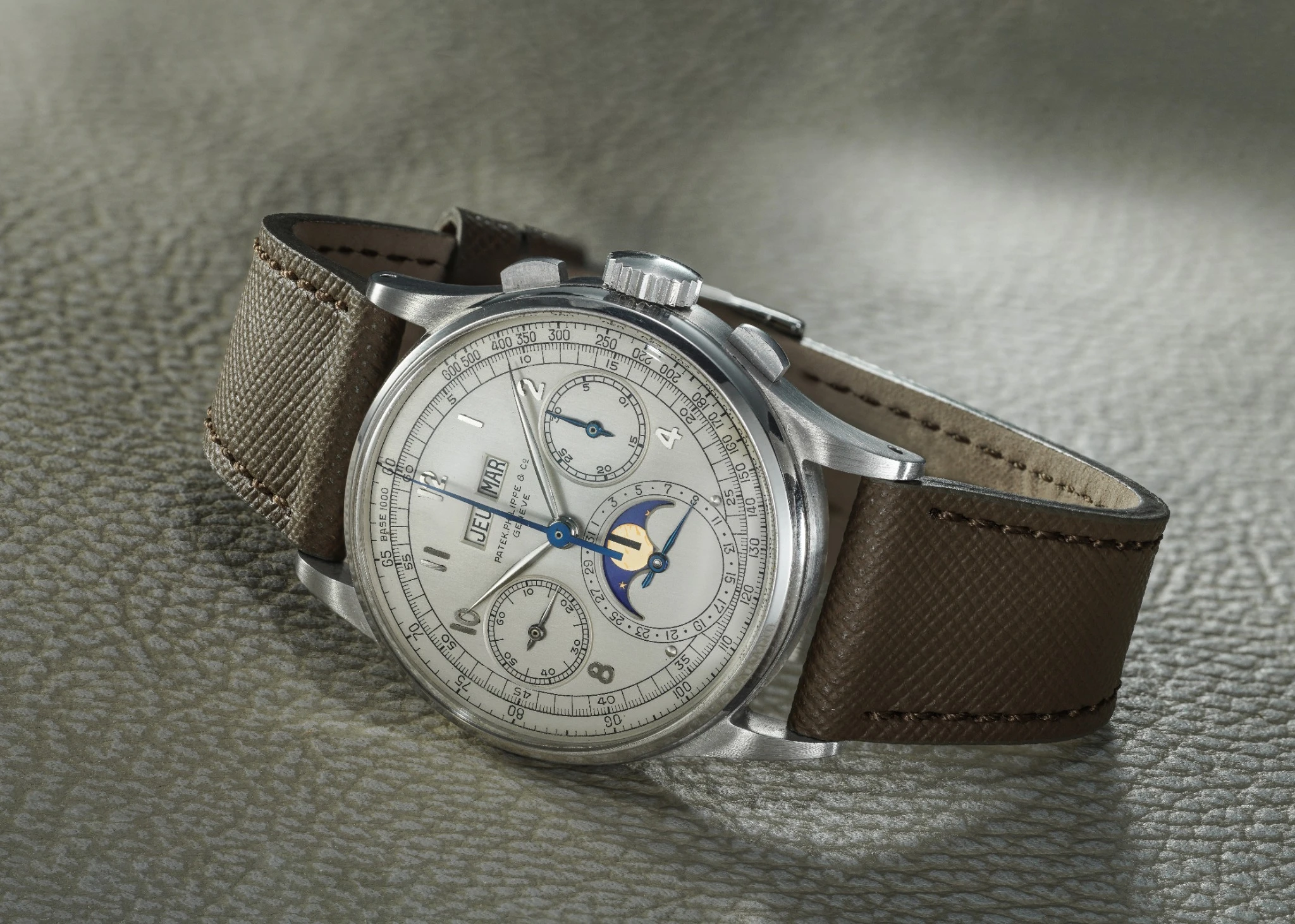 Un orologio da polso di lusso con cassa argentata, quadrante color crema, cinturino in pelle marrone testurizzata e contatori multipli, tra cui un indicatore delle fasi lunari - che riecheggia i pezzi visti all'asta 10 anni Phillips - poggia su una superficie grigia testurizzata.