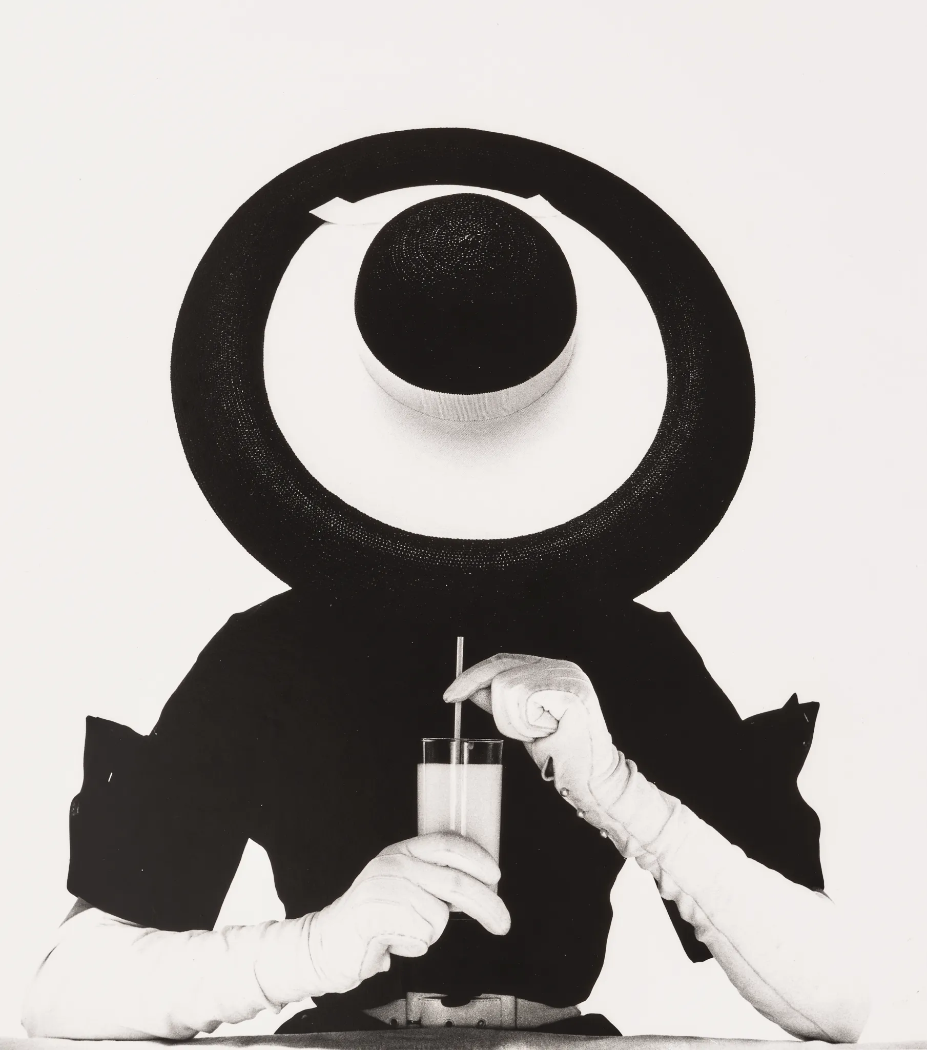 Una persona con un grande cappello circolare e guanti bianchi tiene in mano un bicchiere con una cannuccia. Il volto è oscurato dal cappello, creando un aspetto astratto che ricorda un'asta di Irving Penn. La foto in bianco e nero presenta uno sfondo minimalista.
