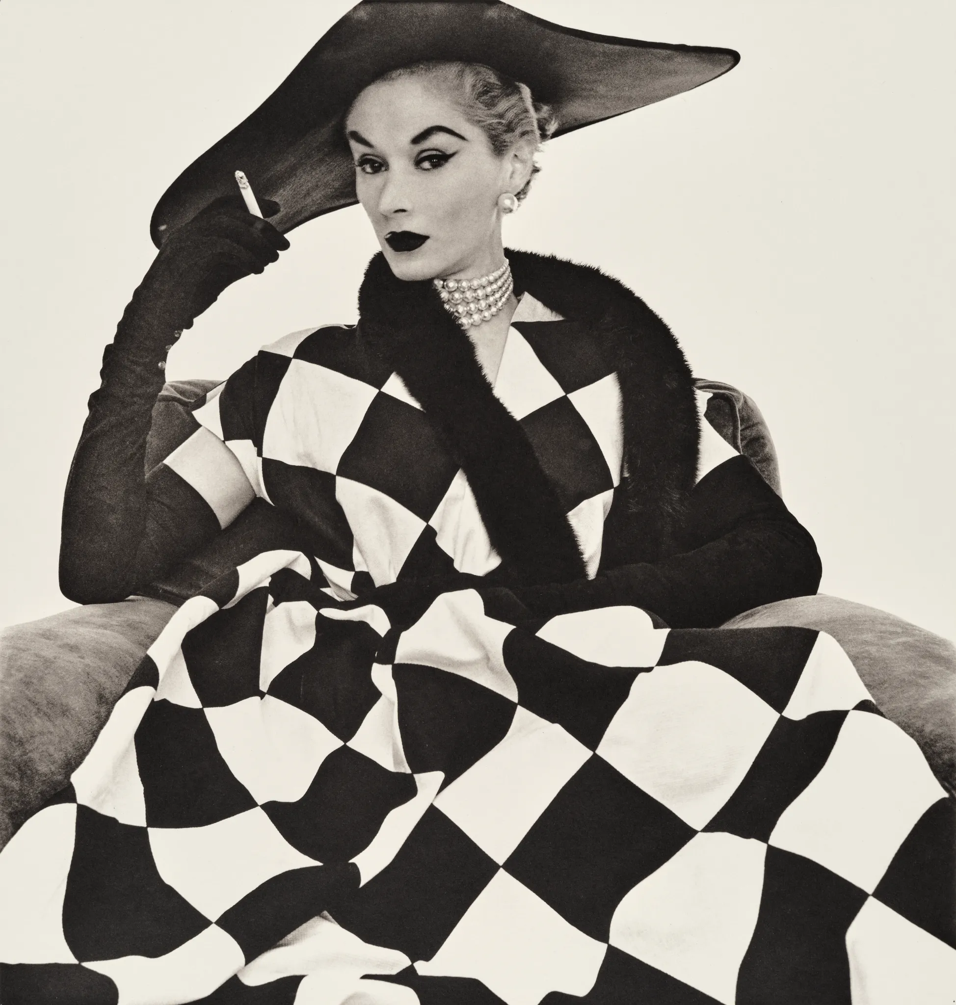 Una donna con un drammatico abito a scacchi e un cappello a tesa larga siede su una sedia e tiene in mano una sigaretta. Con guanti lunghi, una collana di perle e un rossetto scuro, emana un fascino vintage che ricorda una fotografia di Irving Penn.