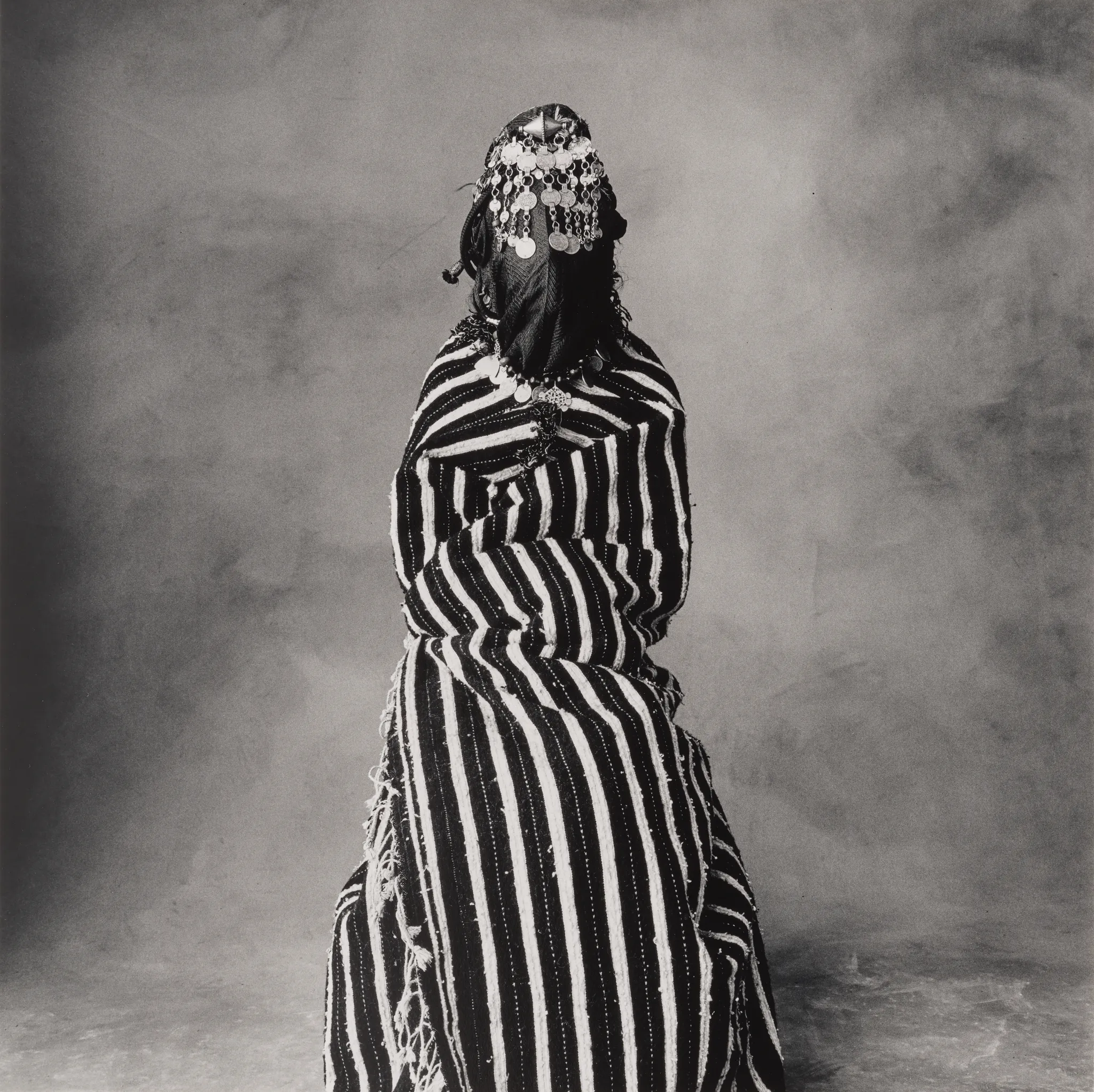 Una persona è seduta di fronte a noi, avvolta in un abito a righe bianche e nere, con gioielli e un copricapo ornato di ornamenti metallici - che richiamano il raffinato stile ritrattistico di Irving Penn - su uno sfondo semplice e strutturato.