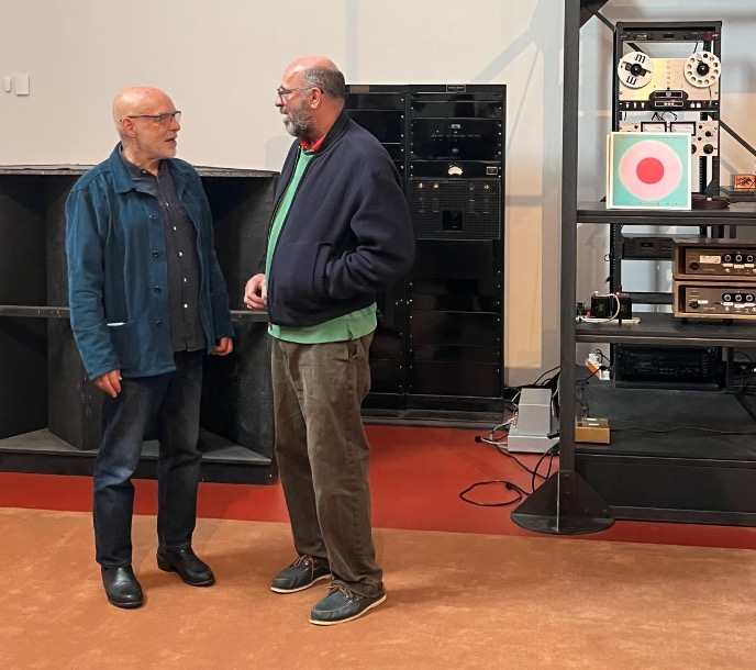 Due uomini stanno in piedi e parlano davanti ad apparecchiature audio, tra cui altoparlanti, macchine a nastro e amplificatori, in una stanza con pavimento marrone rossiccio e pareti chiare, che ricorda le ambientazioni immersive delle mostre di Peter Doig alla Serpentine.