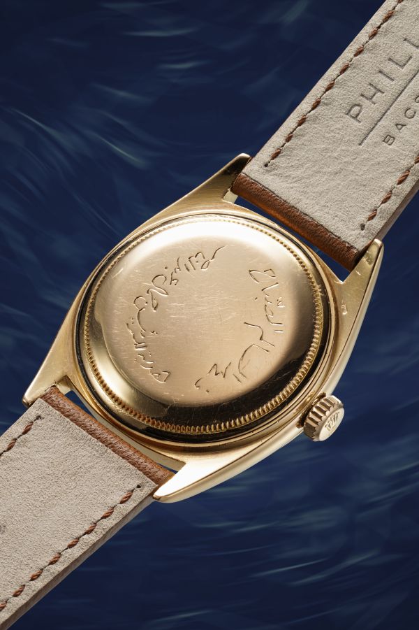 Un orologio da polso d'oro con cinturino in pelle viene mostrato dal retro, rivelando un messaggio inciso in elegante scrittura araba. Lo sfondo è caratterizzato da un motivo blu simile all'acqua.