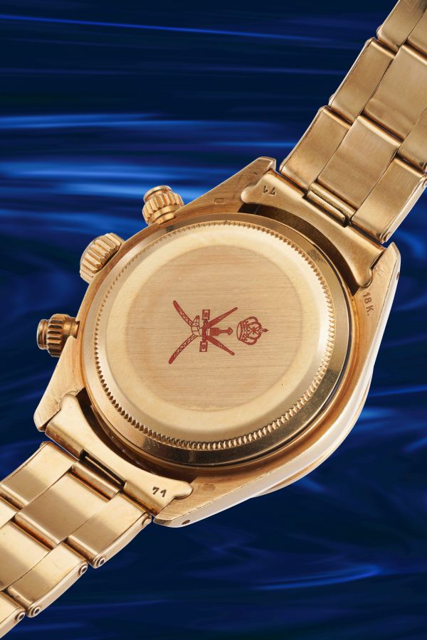 Orologio da polso in oro con emblema rosso inciso con spade incrociate e corona sul retro, su uno sfondo blu vorticoso.